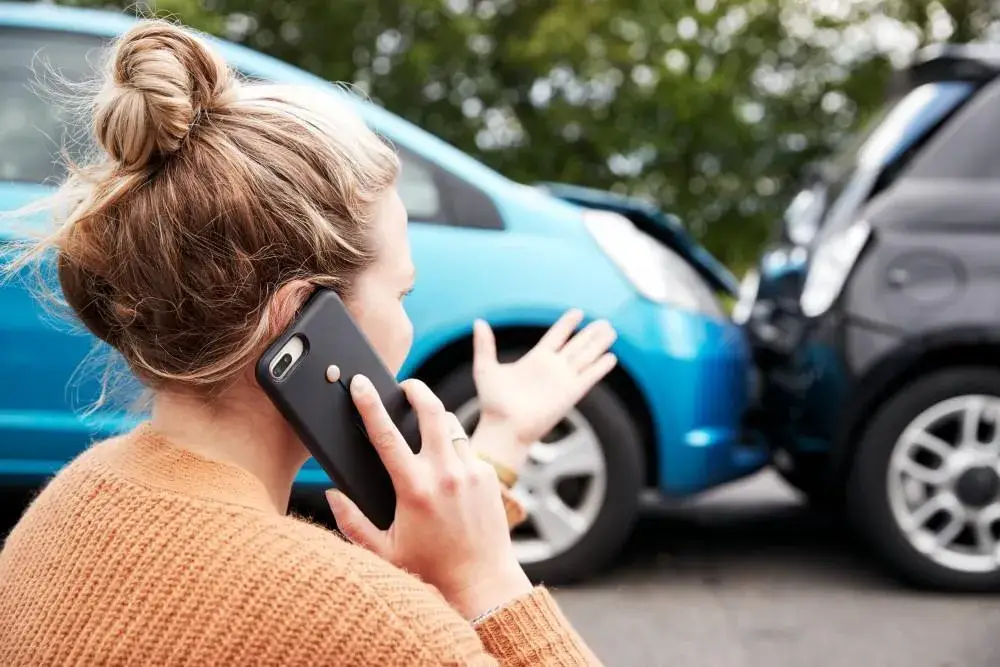 A woman on a phone, seen from behind, with two damaged cars in the background after a collision.
