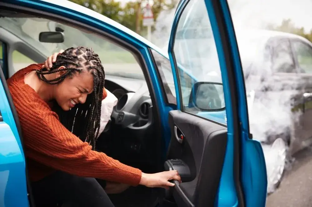 A distressed woman holding her head inside a blue car with a deployed airbag after an accident, with smoke rising from another car in the background.