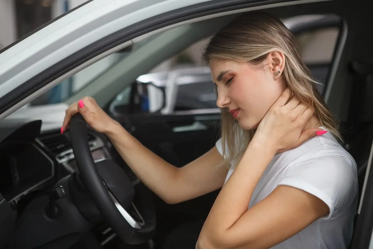 A young woman in a white t-shirt sits in a car, holding her neck with one hand, appearing to be in pain.