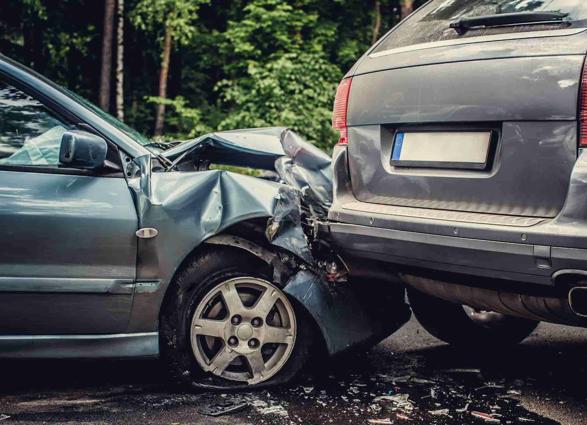 Two cars after a rear-end collision. A light blue sedan has a severely damaged front end, while a gray SUV's rear is visible.