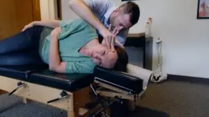 A chiropractor adjusts a female patient's neck as she lies on her side on a chiropractic table.