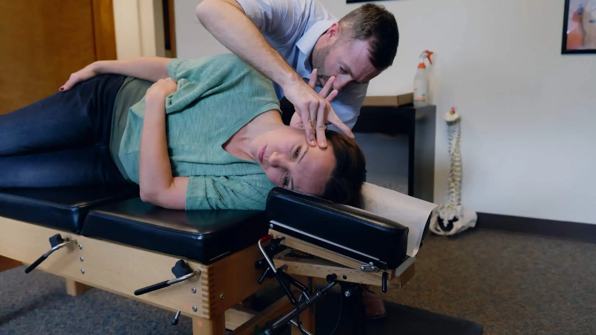 A chiropractor adjusts a female patient's neck as she lies on her side on a chiropractic table.
