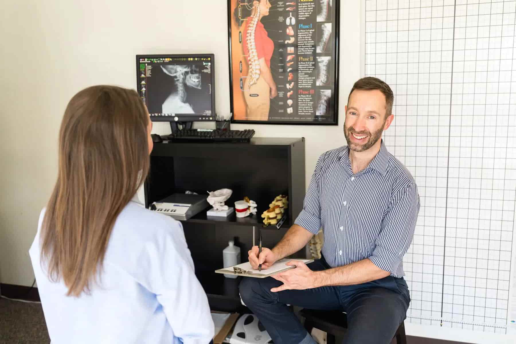 A smiling male chiropractor takes notes on a clipboard while consulting with a female patient whose back is to the camera. A computer monitor shows an X-ray, and anatomical spine models are on a shelf behind them.