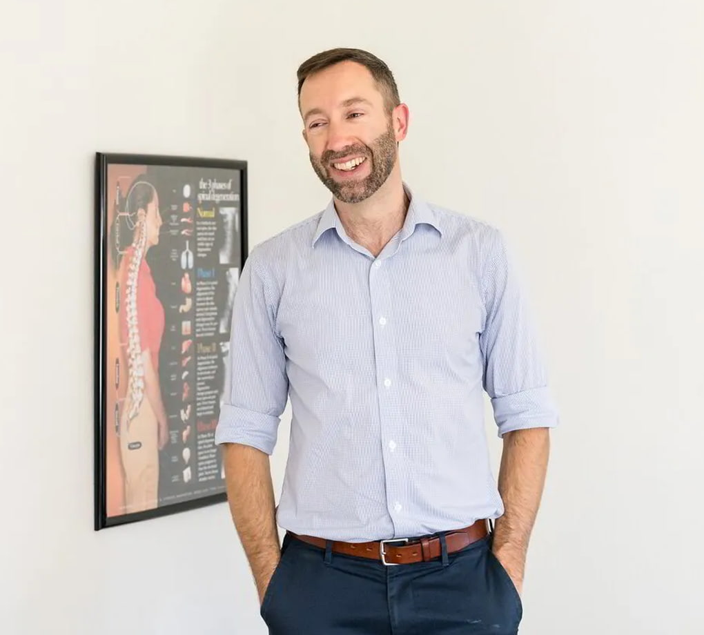 A man with a beard, wearing a light blue shirt and dark pants, smiles while standing in front of a framed poster depicting spinal anatomy.