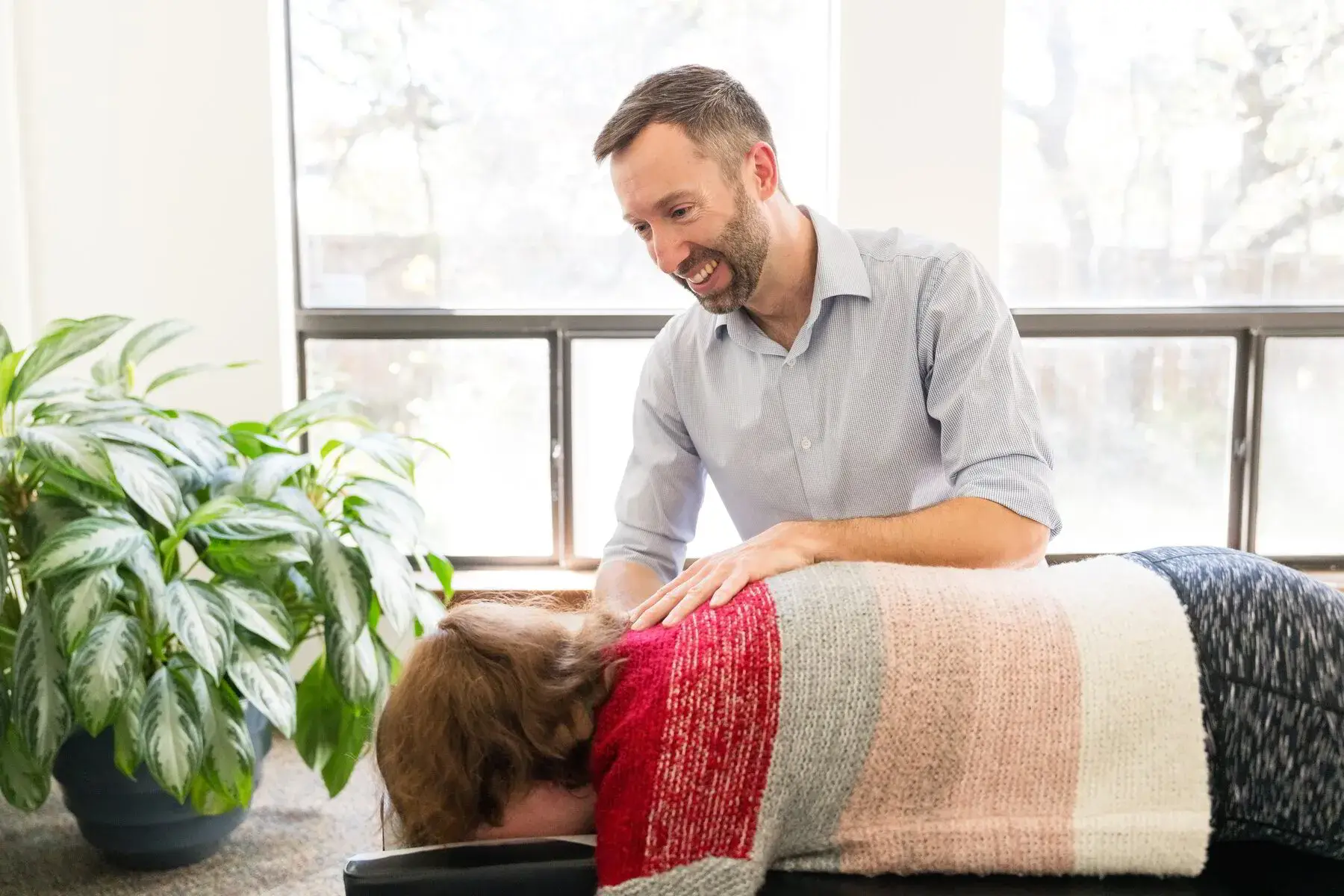 A male chiropractor with a beard smiles as he treats a woman lying face down on a table, placing his hands on her upper back. She wears a striped sweater. A large plant and windows are in the background.