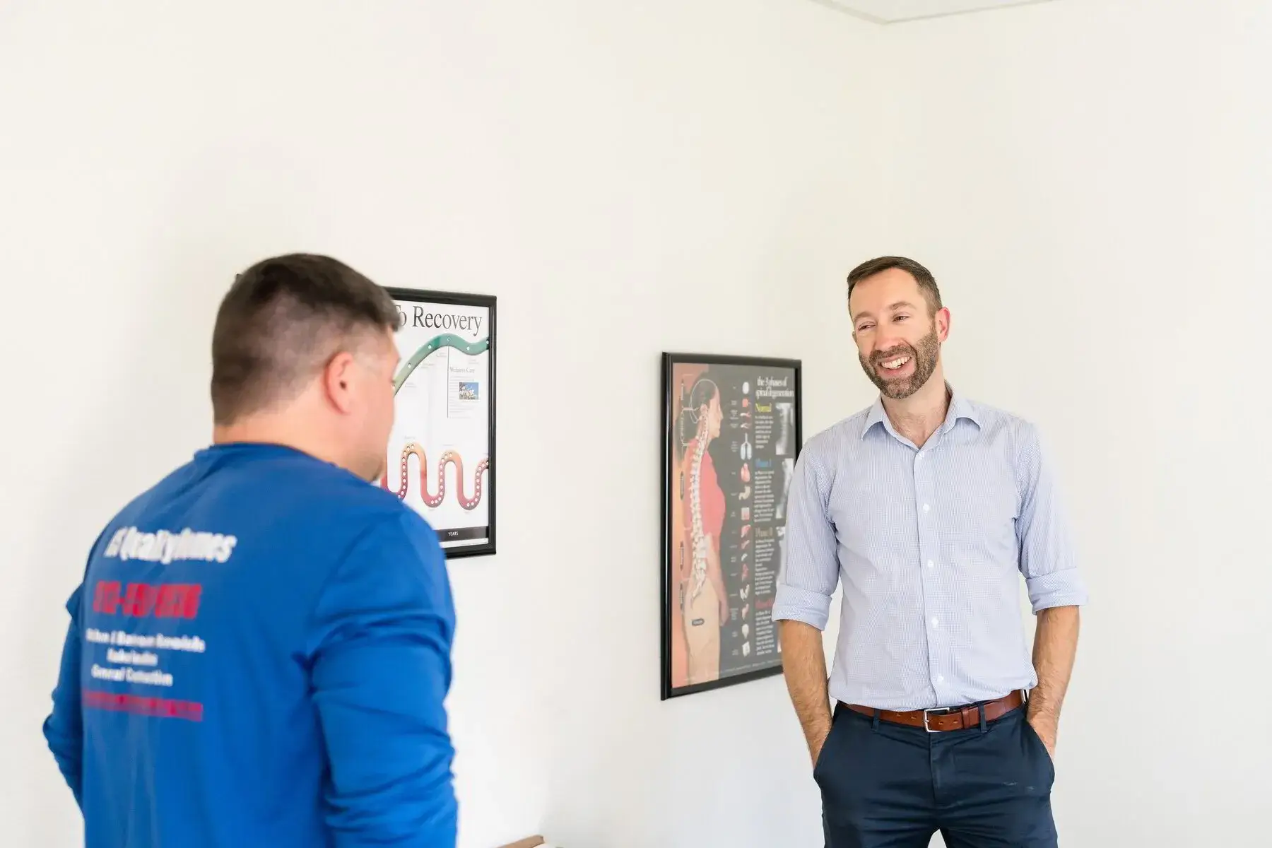 Two men talking in a bright office, one in a blue work shirt, the other in a button-up, with anatomical posters on the wall.