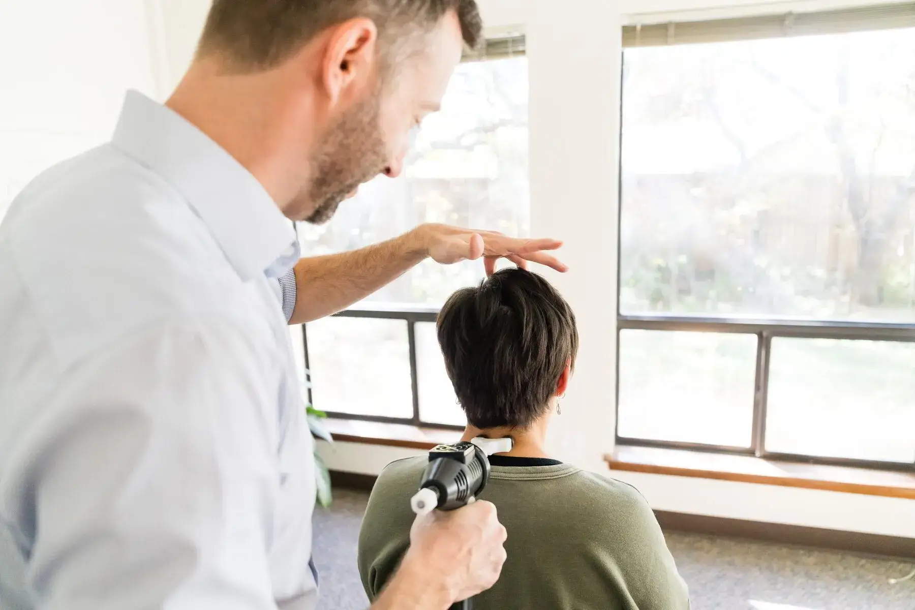 A man uses a handheld activator tool on a woman's upper back/neck, with his other hand above her head. They are in a bright room with large windows.
