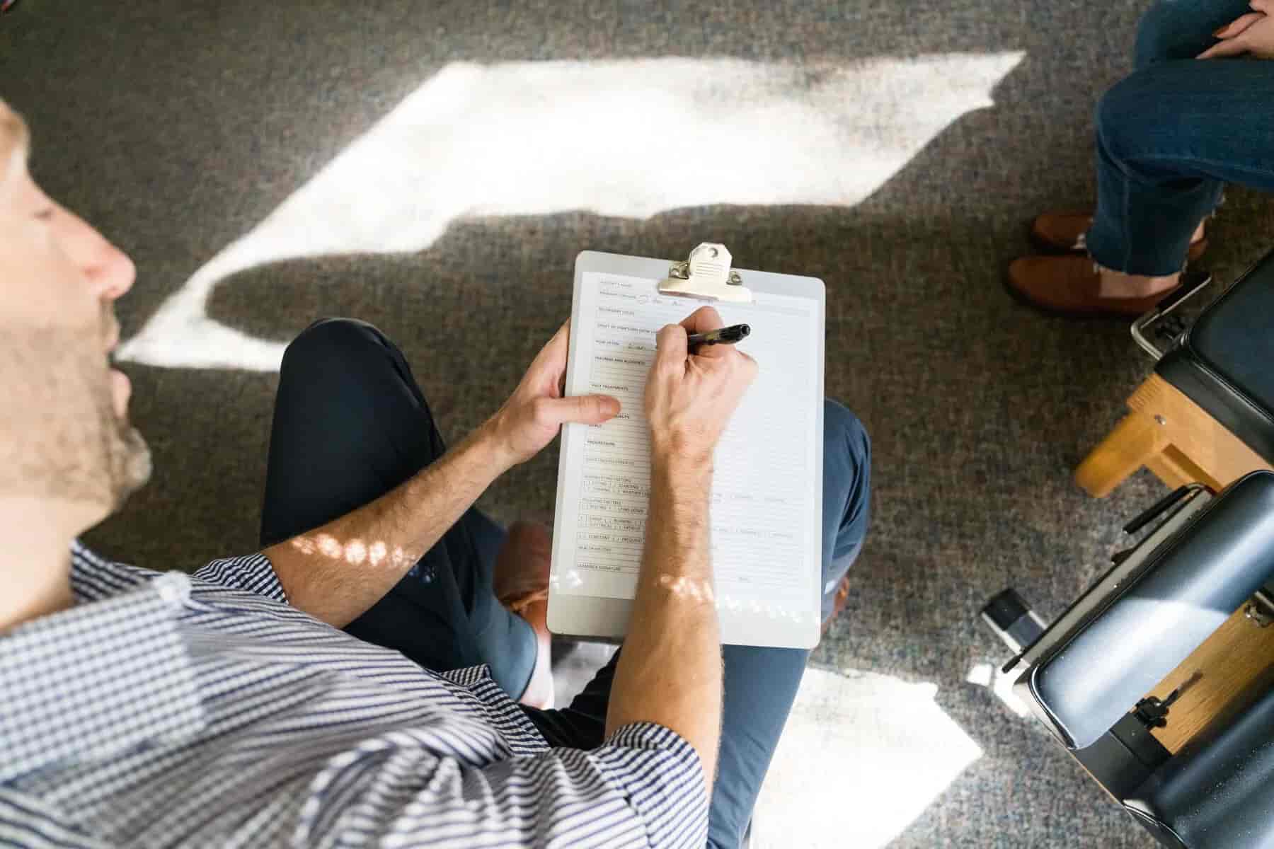 Overhead view of a person with a beard, wearing a striped shirt, filling out a form on a clipboard with a pen. Sunlight shines on the carpet and their arm.