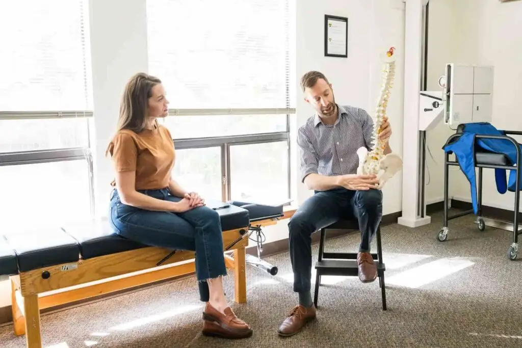 A man in a striped shirt shows a spine model to a woman sitting on an examination table in a medical office.