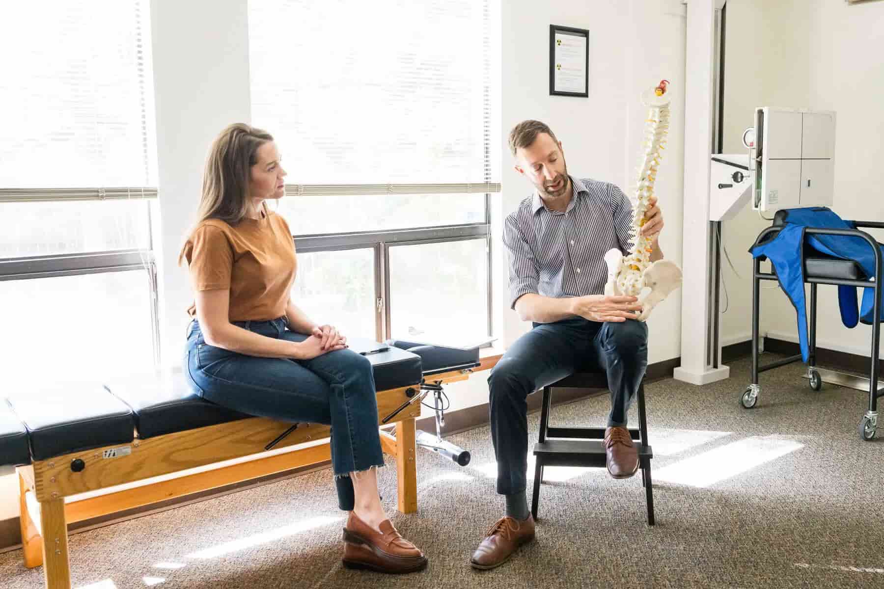 A man in a striped shirt shows a spine model to a woman sitting on an examination table in a medical office.