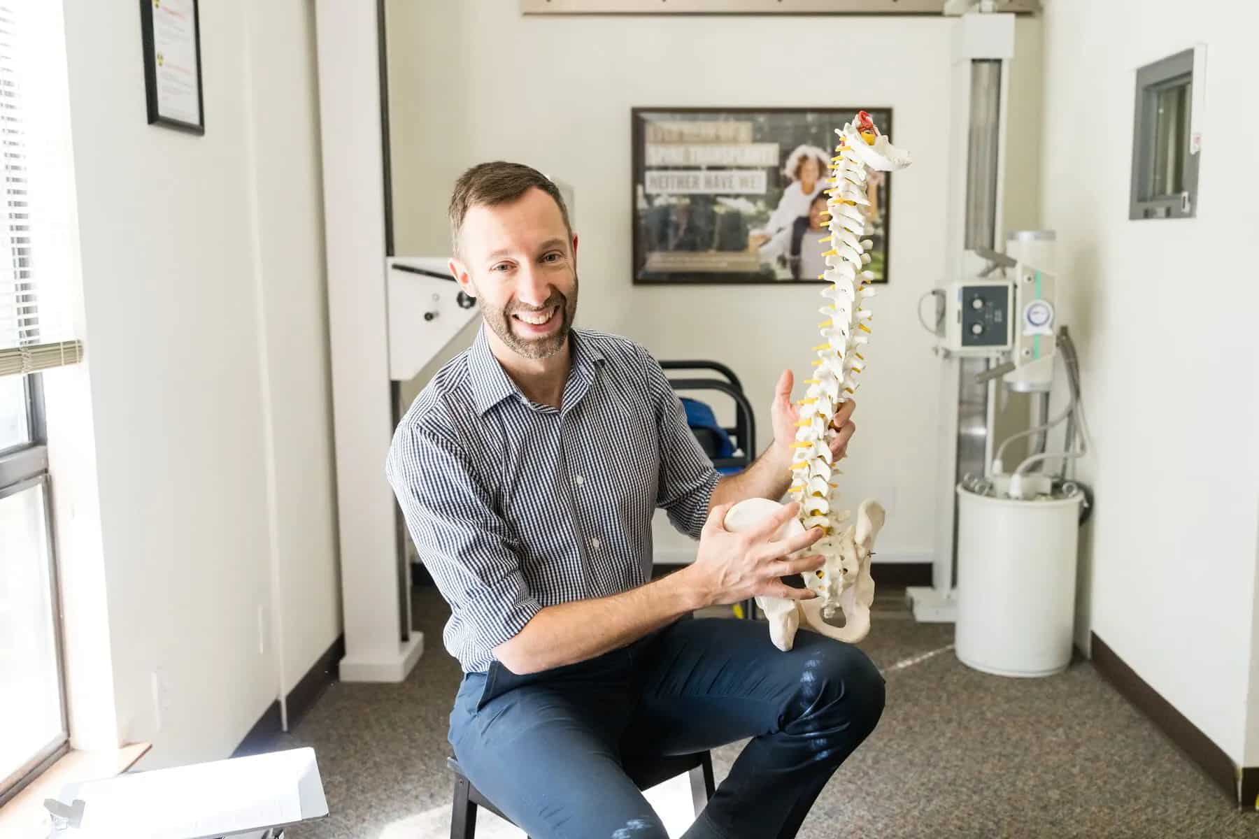 A smiling man in a striped shirt holds a human spine model in a medical office, with medical equipment and a window in the background.