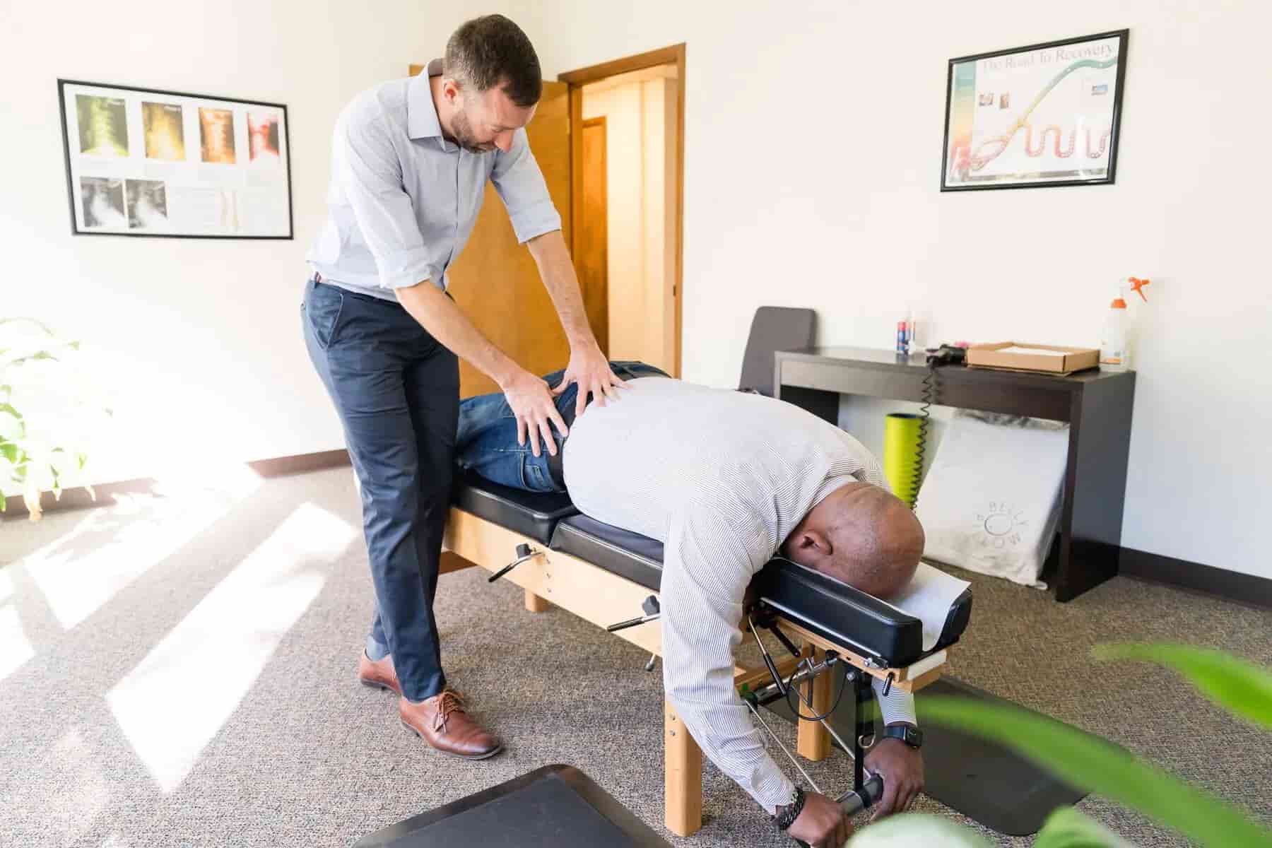 A chiropractor adjusts a patient's lower back on an adjustment table in a bright office.