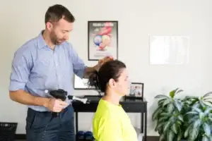 A man in a blue shirt uses a handheld therapeutic device on a woman's neck, who is wearing a yellow shirt and has her eyes closed.