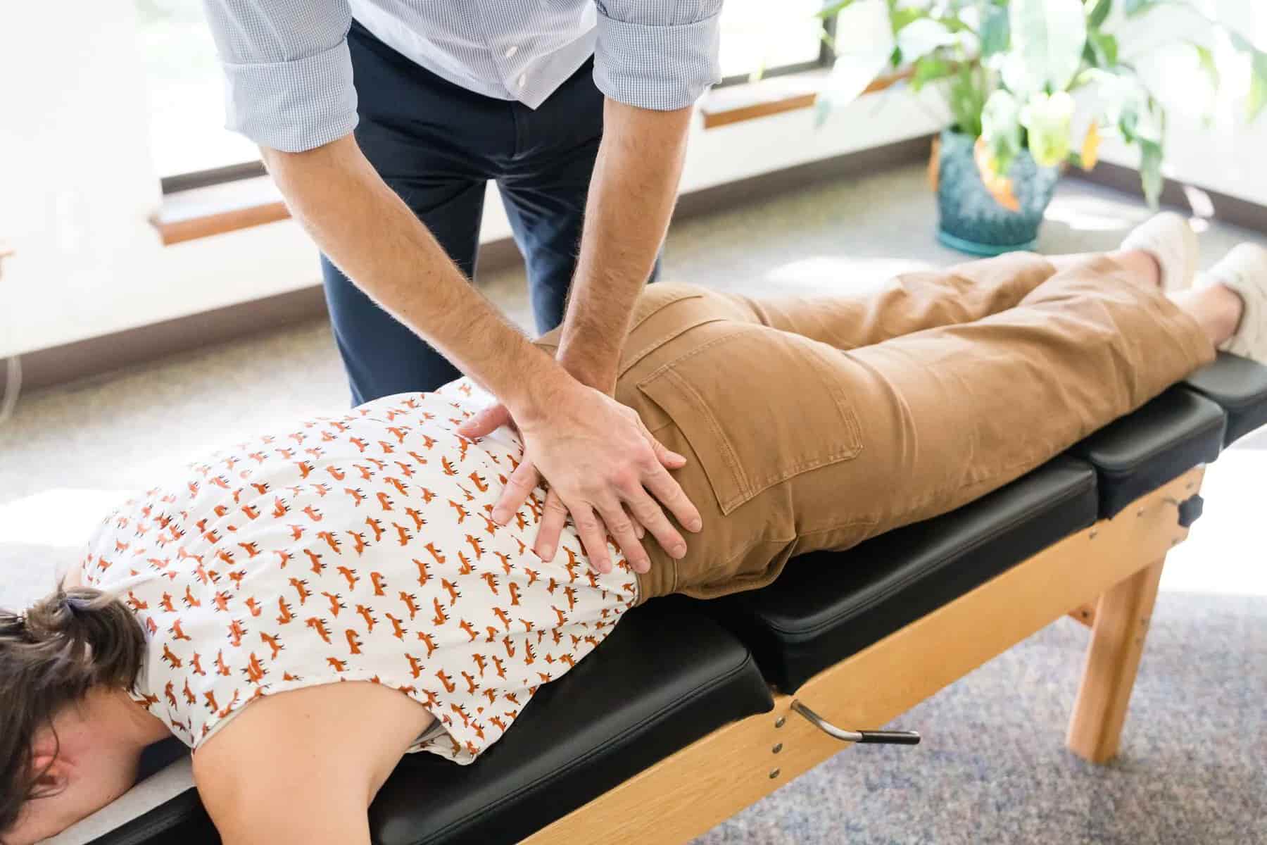 A chiropractor adjusts a patient's lower back on a chiropractic table. The patient is lying face down, wearing a fox-patterned shirt and tan pants.