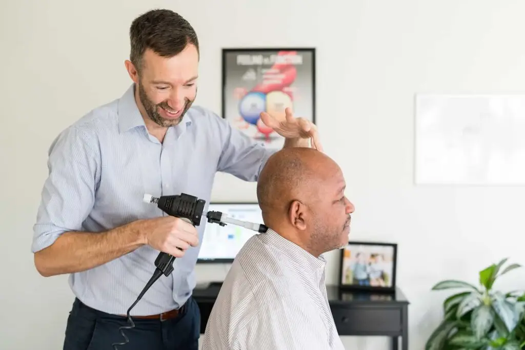 A chiropractor uses a handheld adjustment tool on a patient's neck, while the patient sits facing away.
