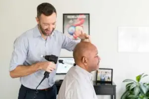 A chiropractor uses a handheld adjustment tool on a patient's neck, while the patient sits facing away.