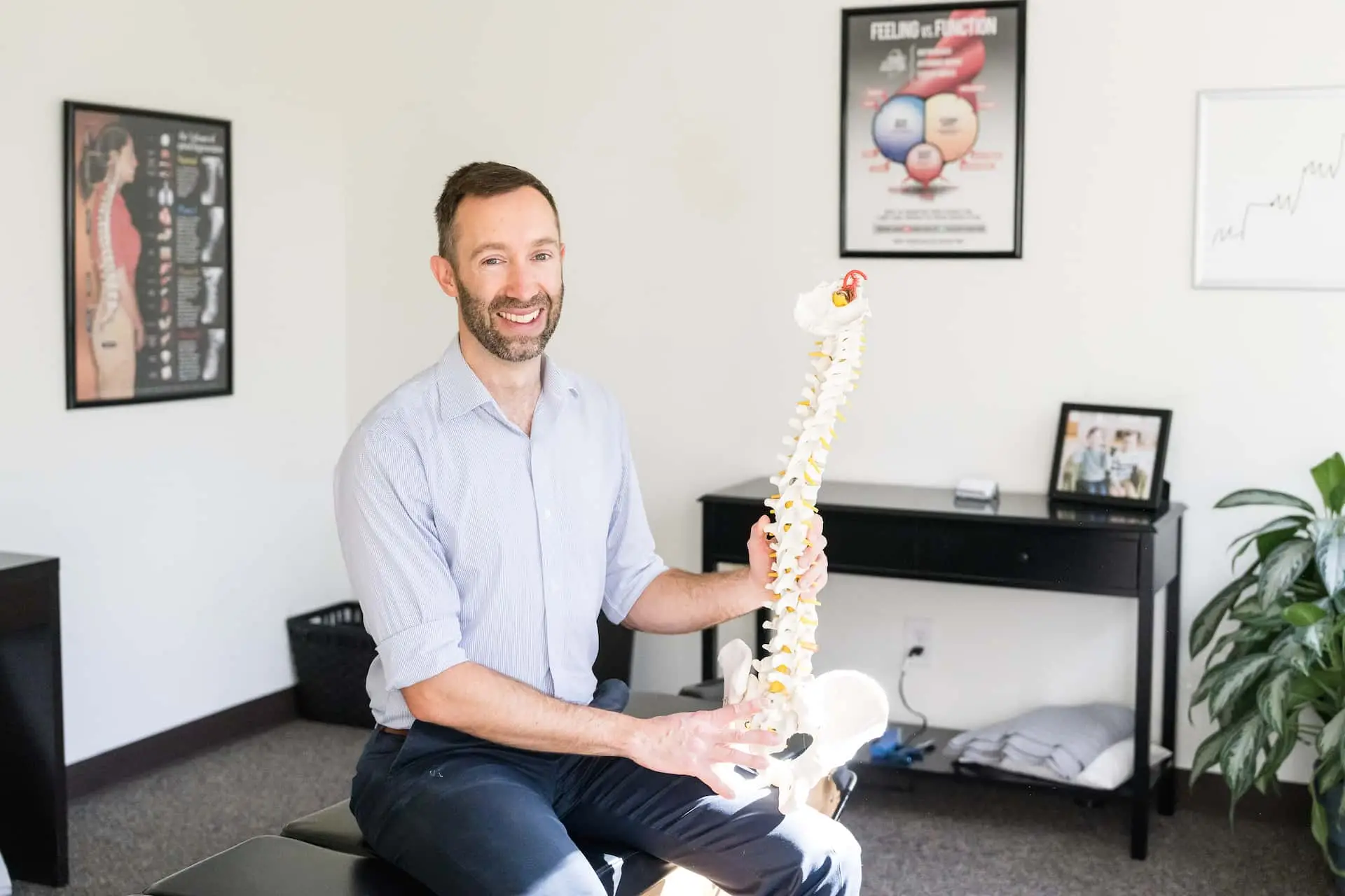 A smiling man, likely a chiropractor, sits on an exam table holding a human spine model in an office setting.