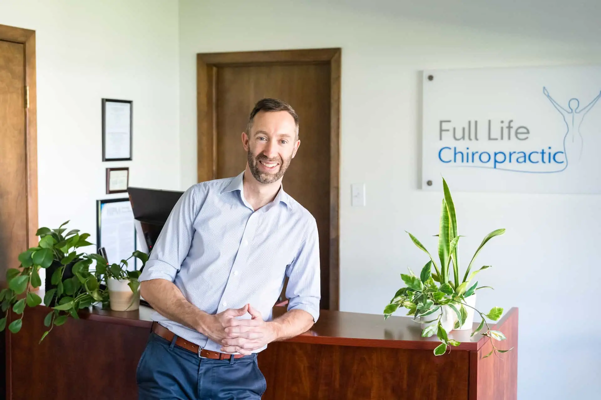 A smiling man in a light blue shirt leans on a dark wooden reception desk at Full Life Chiropractic.