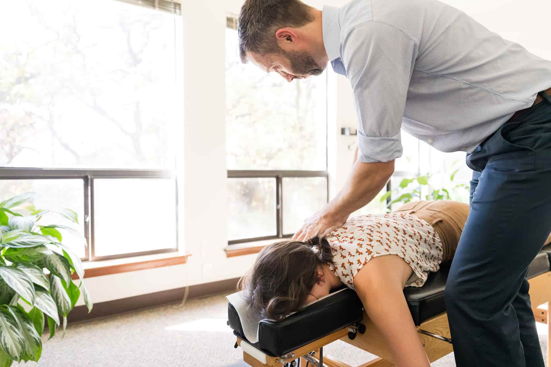 A chiropractor performs an adjustment on a woman's upper back as she lies face down on a treatment table.