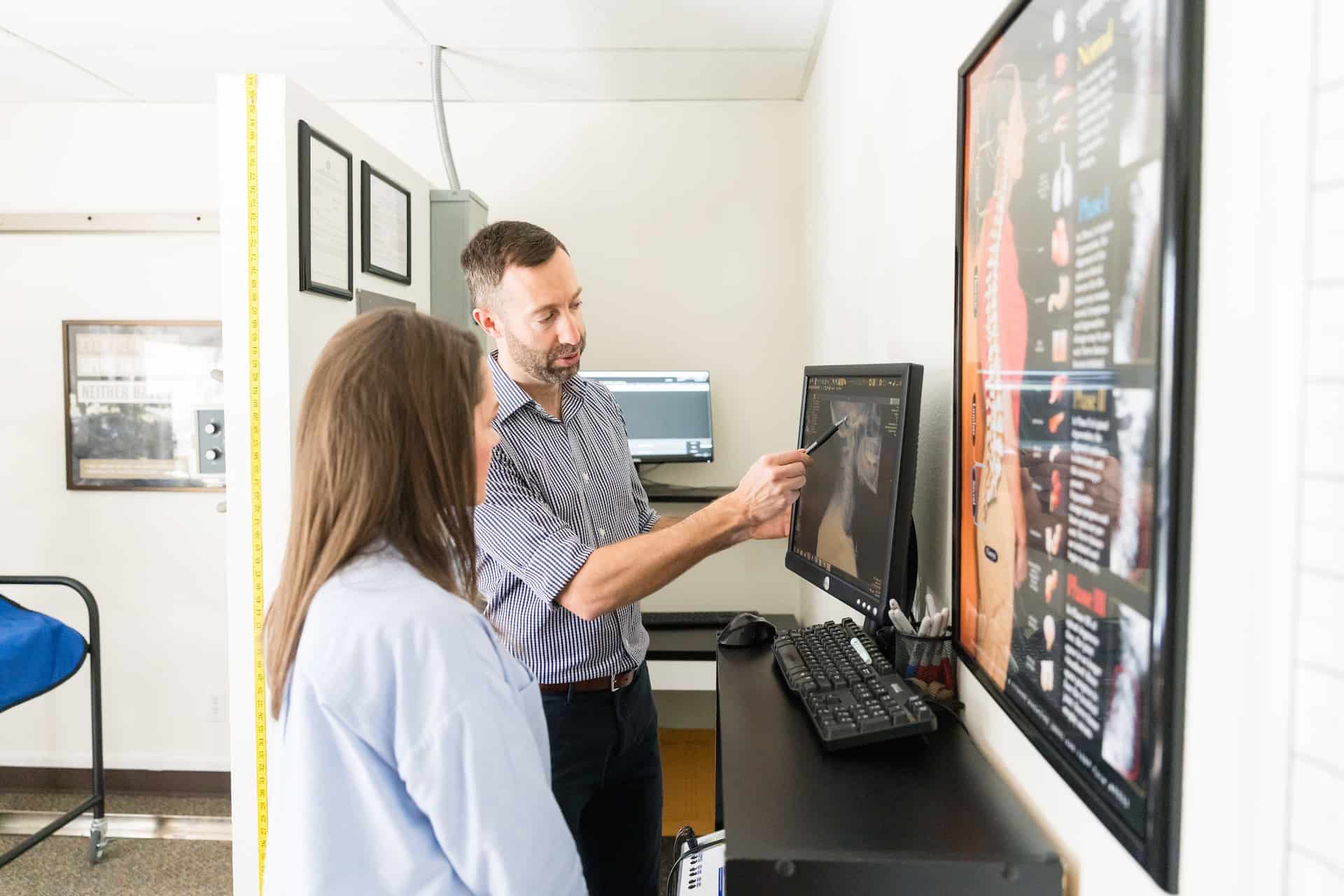 A male healthcare professional points at a computer screen displaying medical imaging while explaining it to a female patient in a clinic.