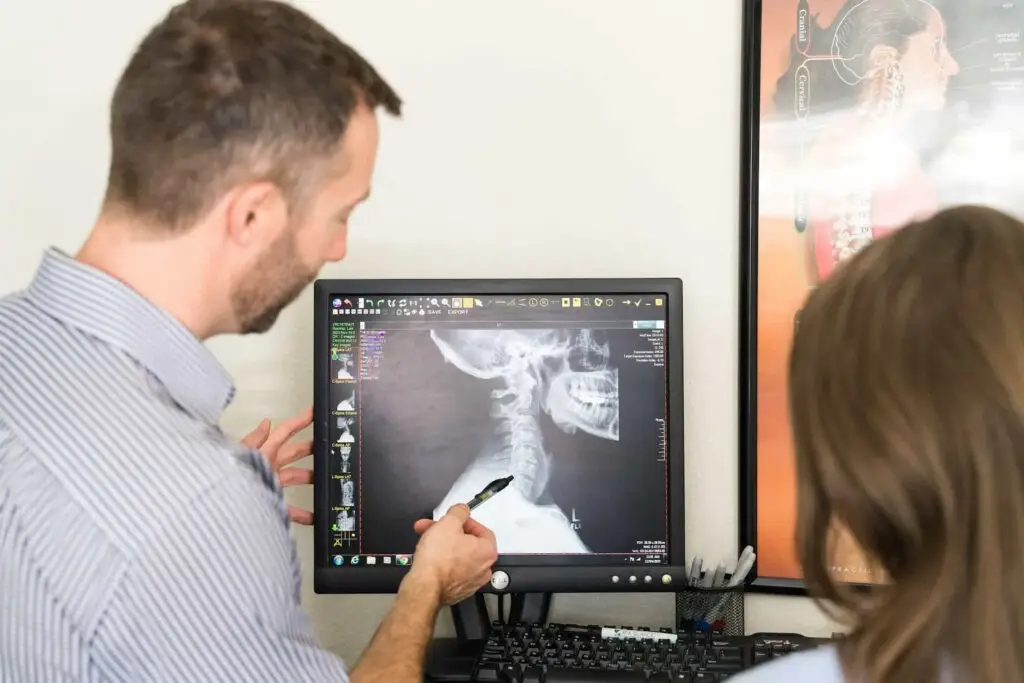 A man points at a cervical spine X-ray on a computer monitor, explaining it to a woman. An anatomical chart is visible in the background.