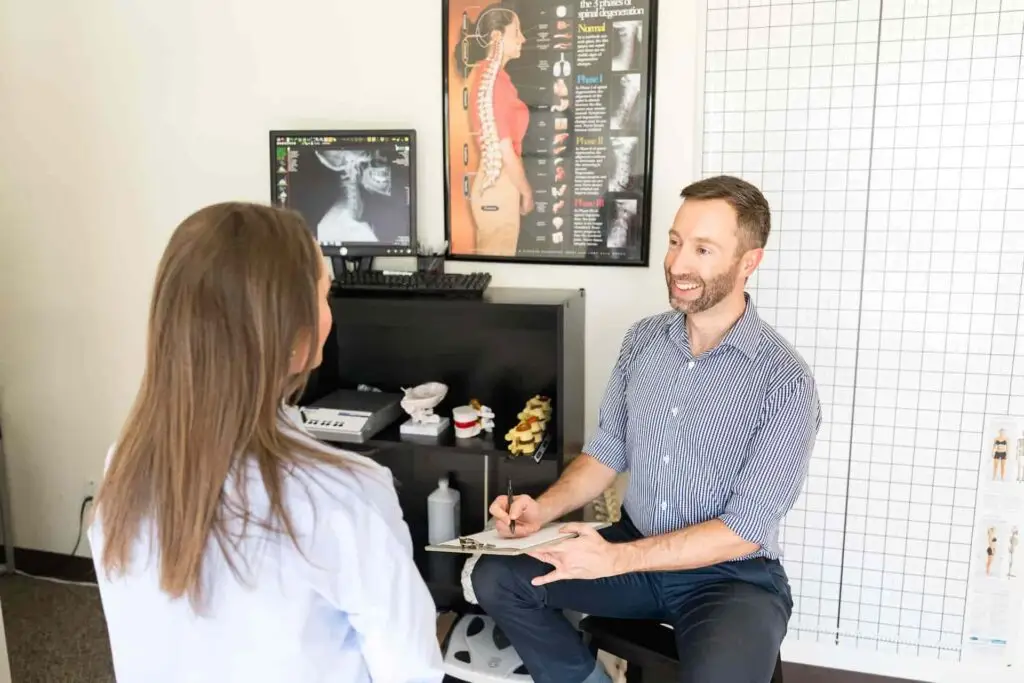 A man and woman in a chiropractic office. The man, a chiropractor, smiles while holding a clipboard, facing the woman whose back is to the camera. An X-ray and spinal charts are in the background.