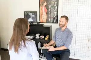 A man and woman in a chiropractic office. The man, a chiropractor, smiles while holding a clipboard, facing the woman whose back is to the camera. An X-ray and spinal charts are in the background.