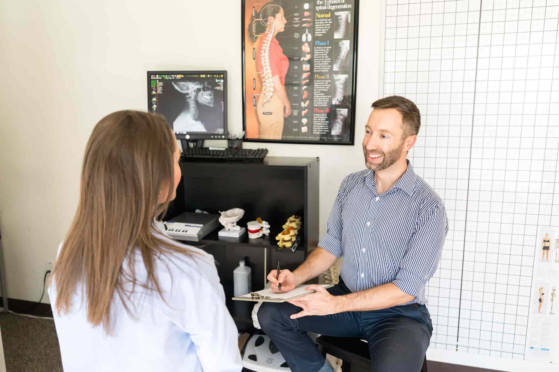 A man and woman in a chiropractic office. The man, a chiropractor, smiles while holding a clipboard, facing the woman whose back is to the camera. An X-ray and spinal charts are in the background.