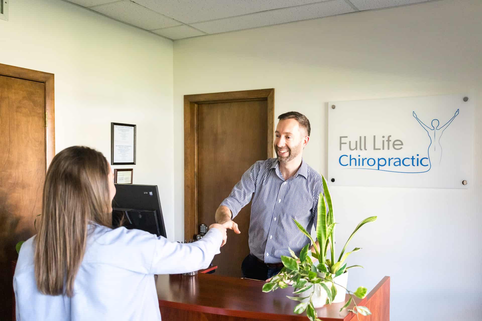A man and woman shaking hands over a wooden reception desk in an office with a "Full Life Chiropractic" sign on the wall.