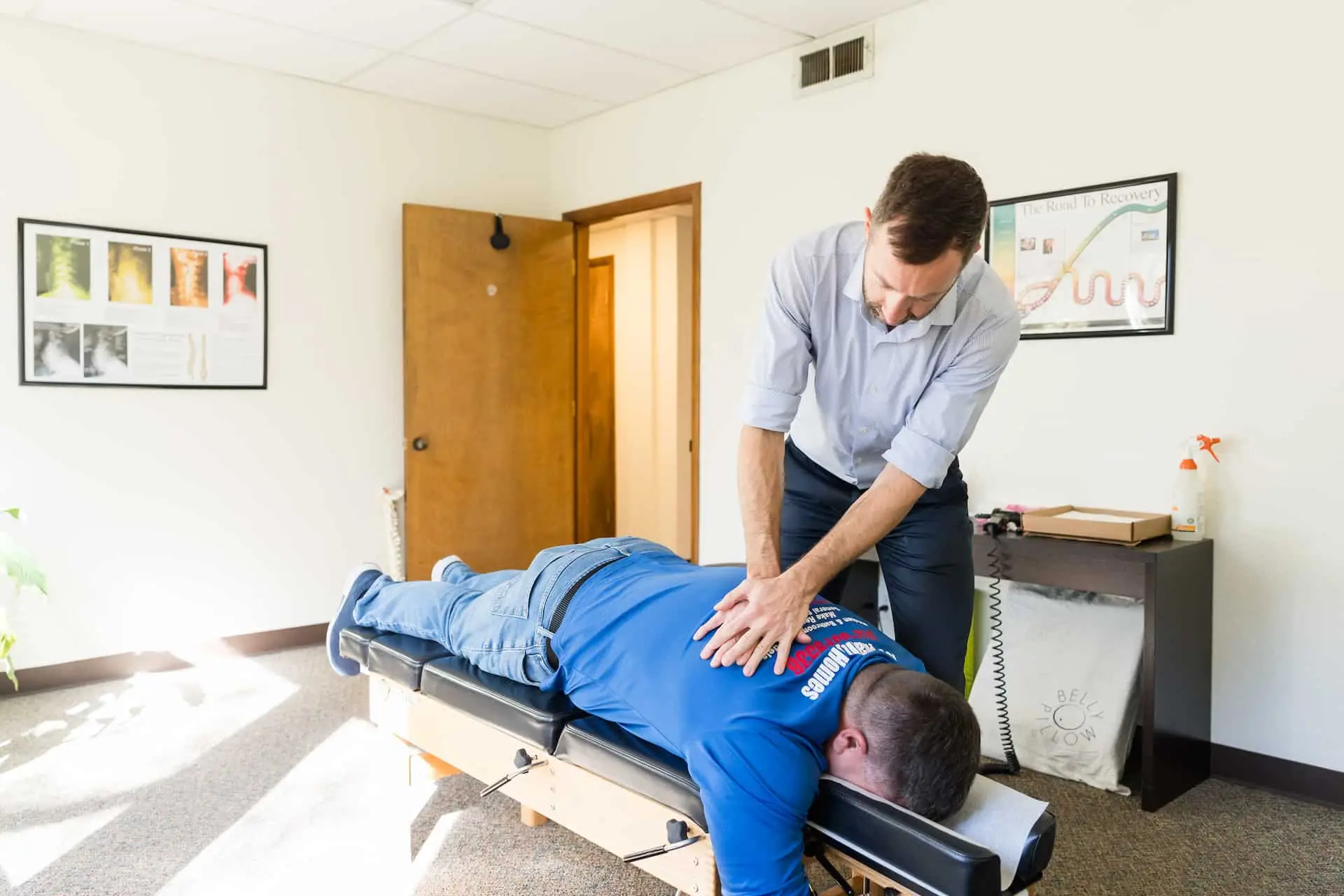 A chiropractor adjusts a male patient's back who is lying face down on an adjustment table in a bright office.