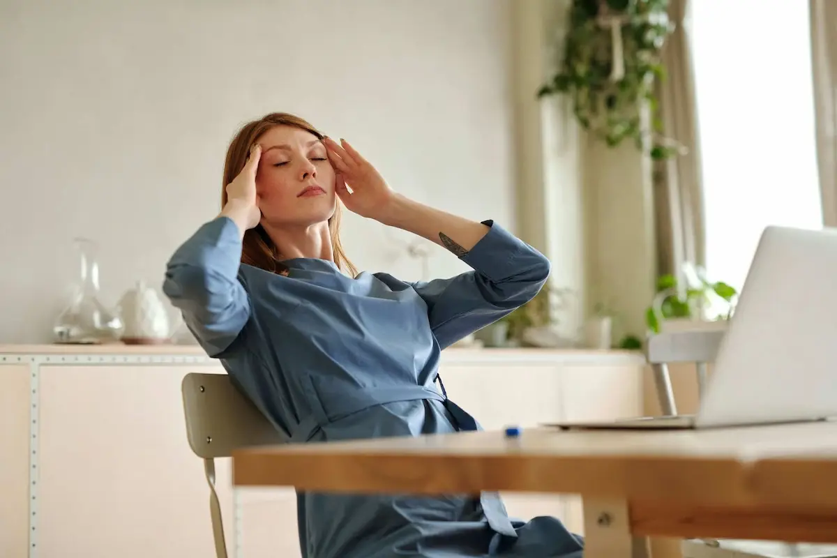 A woman with red hair and freckles, wearing a blue dress, sits with her eyes closed and hands pressed to her temples, appearing to have a headache. A laptop is on the table beside her.