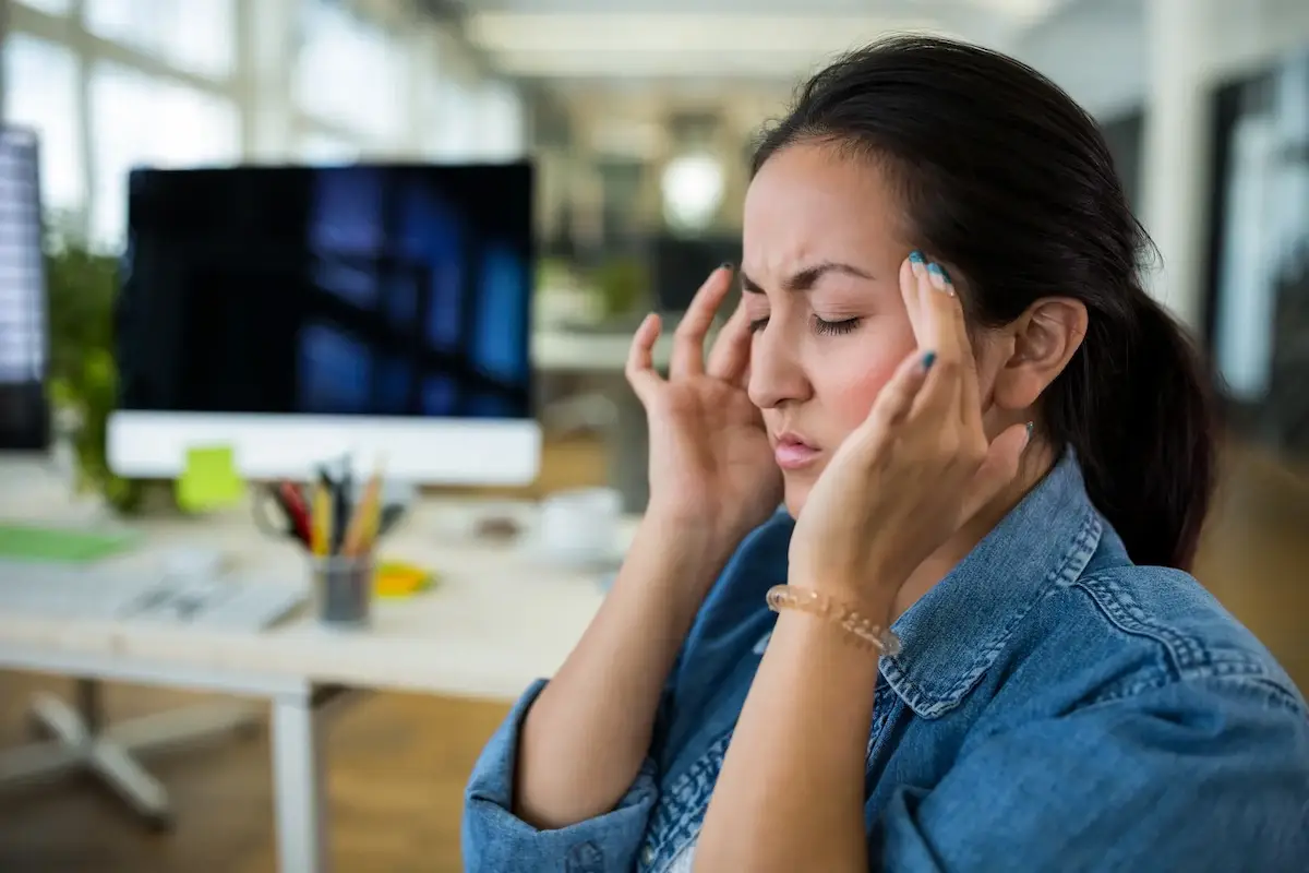 A woman with her eyes closed, holding her temples, indicating a headache or stress, in an office setting.
