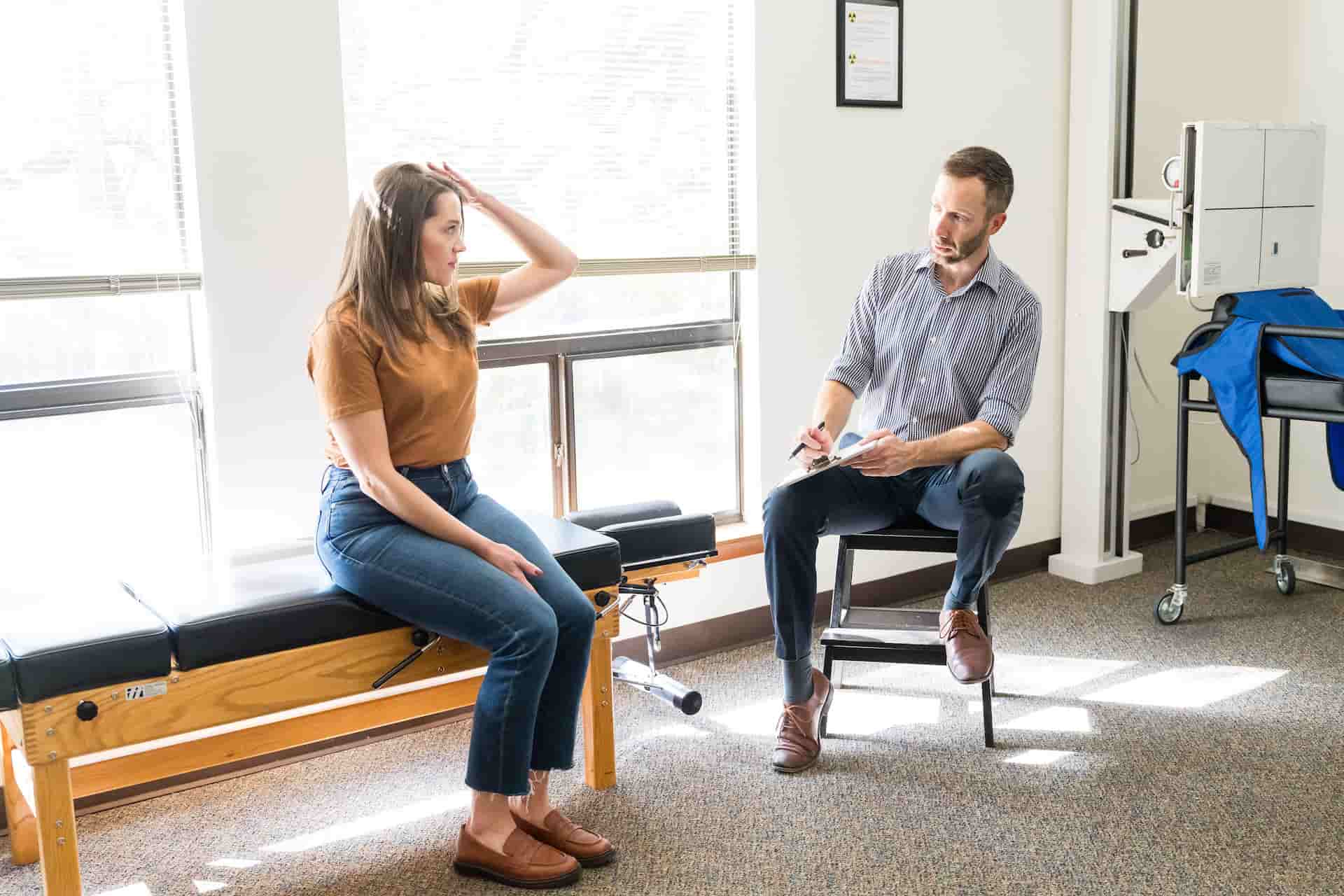 A woman sitting on an examination table with her hand to her head, talking to a man with a clipboard in a medical office.