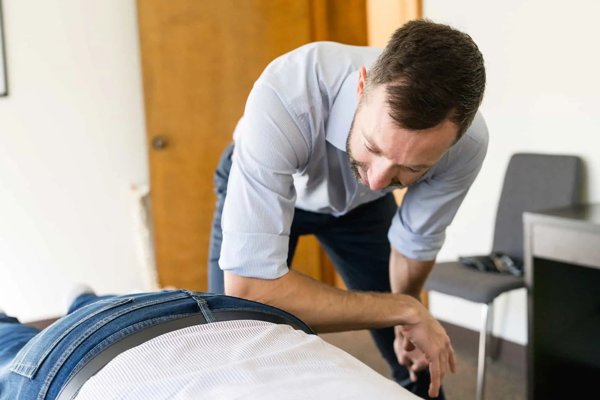 A man in a light blue shirt leans over a person lying face down on a table, performing a treatment or adjustment.