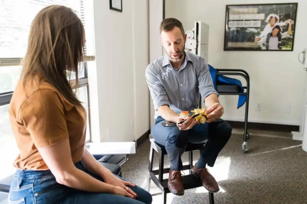 A man in a striped shirt explains a spine model to a woman in a brown shirt and jeans in an office setting.