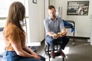 A man in a striped shirt explains a spine model to a woman in a brown shirt and jeans in an office setting.