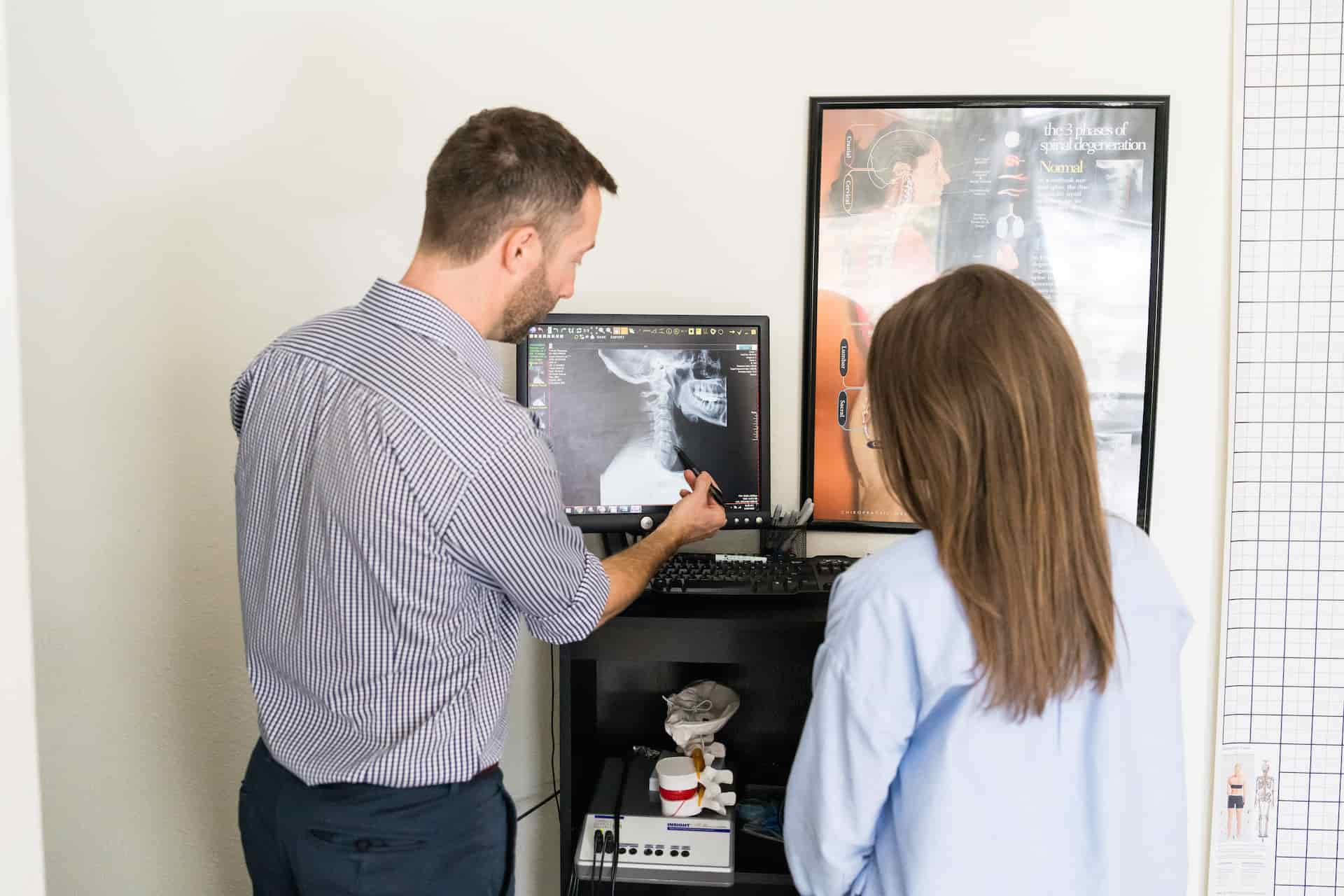 A man in a striped shirt points at a neck X-ray on a computer screen while a woman in a blue shirt looks on in a medical office.