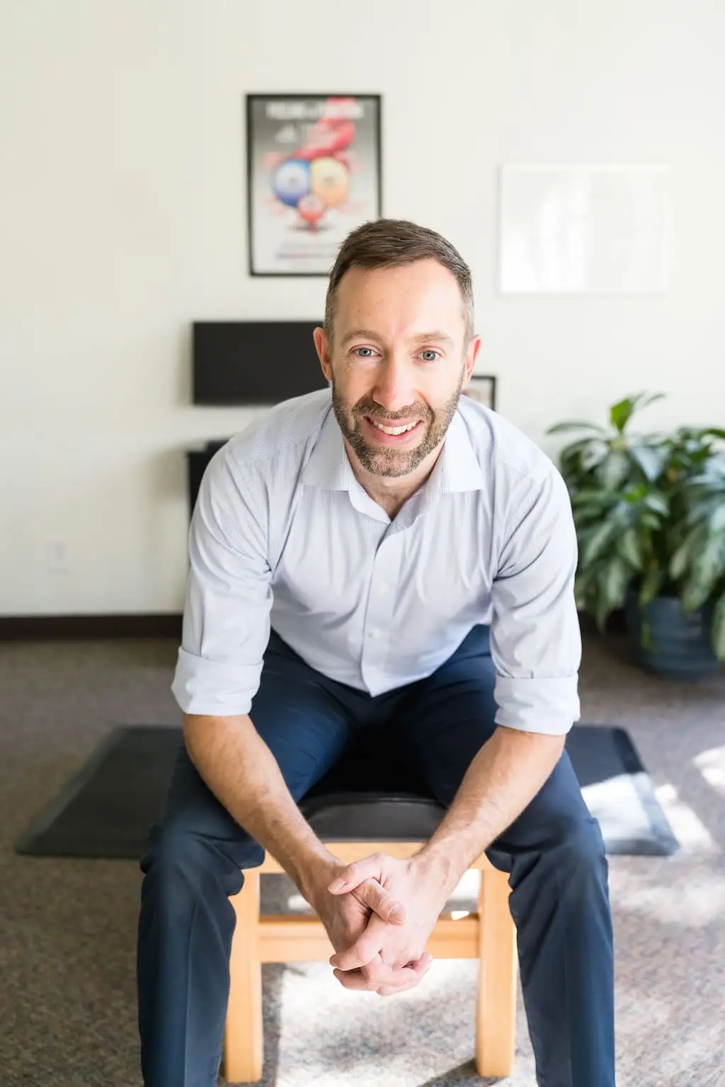 A smiling man with a beard and blue eyes sits on a stool, wearing a light blue shirt and dark pants, in a bright room with a poster and plant in the background.