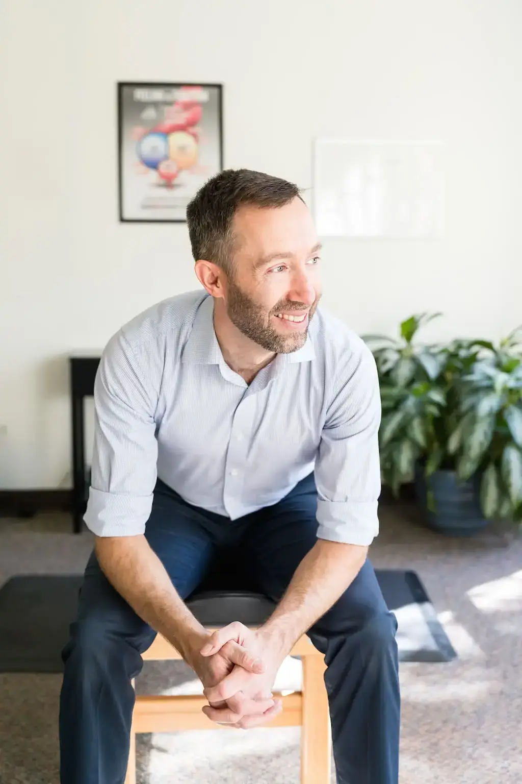 A smiling man with a beard, wearing a light blue shirt, sits on a wooden stool, looking right.