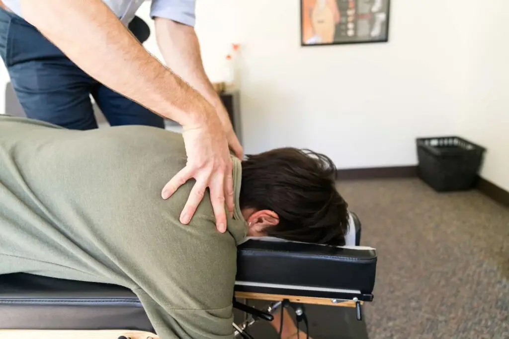 A chiropractor adjusts a patient's upper back and shoulder while they lie face down on an examination table.