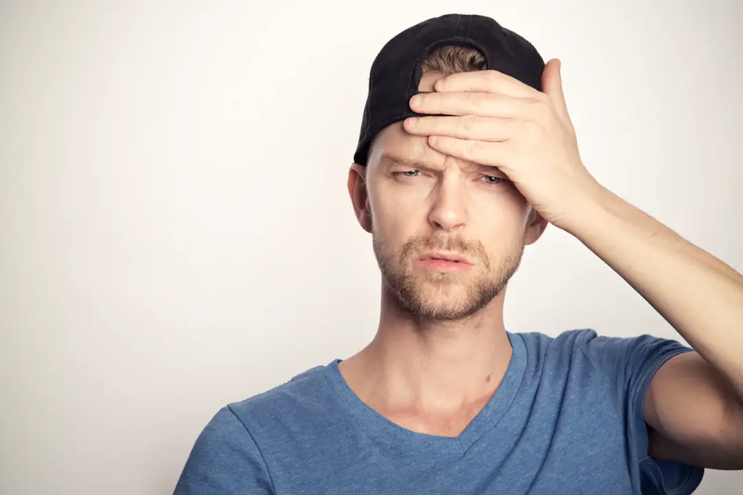 A man with a black cap and blue shirt, hand on forehead, looking confused or in pain.
