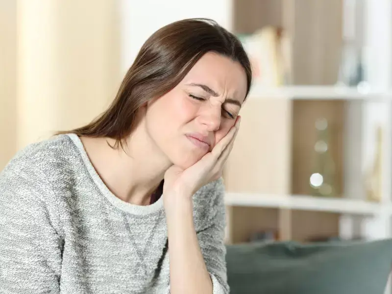 A woman with a pained expression, eyes closed, holding her right cheek with her hand, indicating toothache or jaw pain.